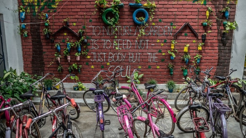 Around ten colorful bikes are parked in front of a red brick wall with plants hanging from the wall. On the wall in white capital letters is written:"To nurture a garden is to feed not just body but the soul." The wal lis deorated with plants in colorful tires or pots.